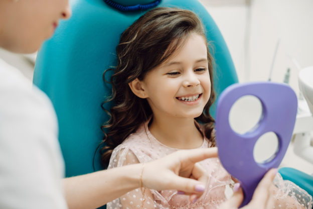 Portrait of a cute little girl looking at her teeth after doing teeth surgery in a pediatric stomatology while sitting in stomatology seat.